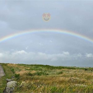 Full rainbow over the Inverness boardwalk and town in Cape Breton, Nova Scotia, with three smiling emojis with heart eyes, star eyes, sunglasses floating over the rainbow.